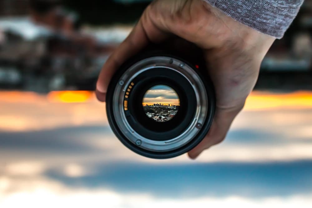 A removable camera lens is held so that we can see a blurry landscape background in crispo focus
